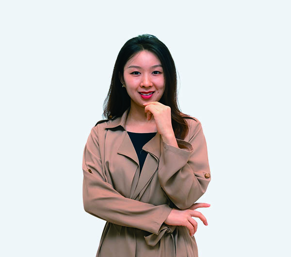 A woman in a beige trench coat smiling at the camera with one hand under her chin.