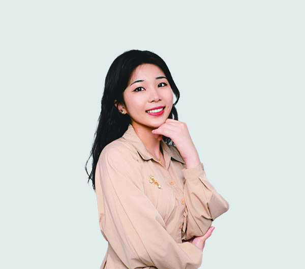 A smiling woman with black hair, wearing a beige blouse, posing with one hand on her chin against a light background.
